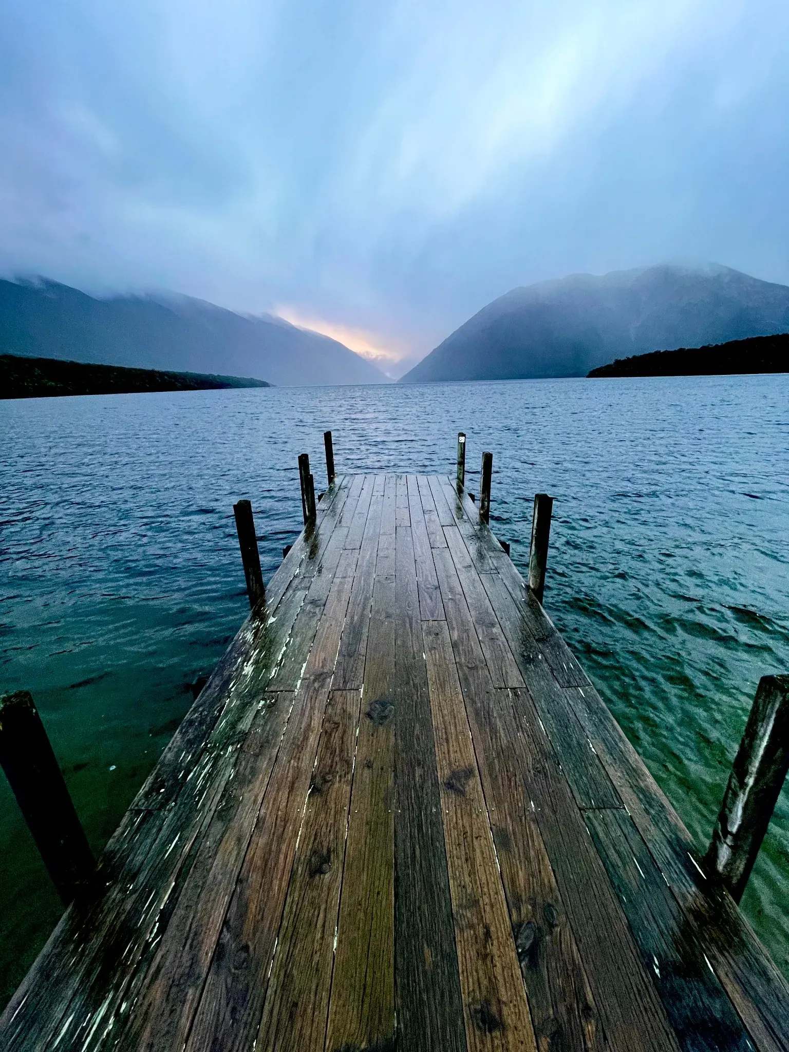 A wooden jetty reaching into still water beneath mountains at dawn